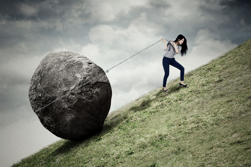 Image of young businesswoman climbing on the hill while pulling big stone with a chain