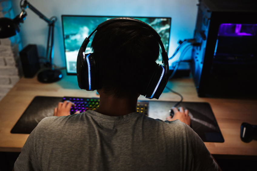 Back view of teenage gamer boy playing video games online on computer in dark room wearing headphones with microphone and using backlit colorful keyboard